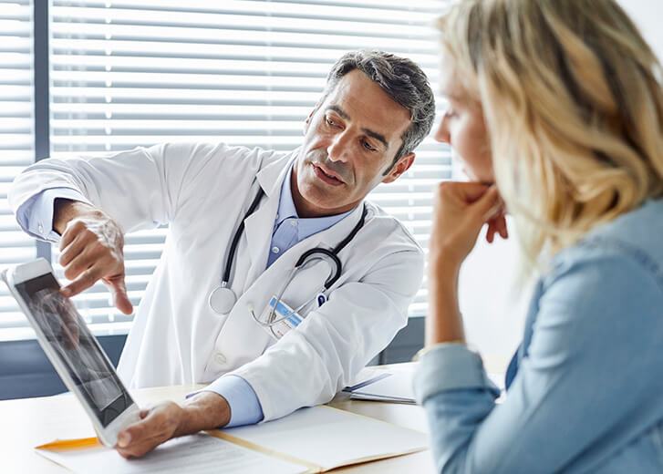 Doctor showing digital tablet to woman. Female patient sitting with health professional. They are against window in hospital.