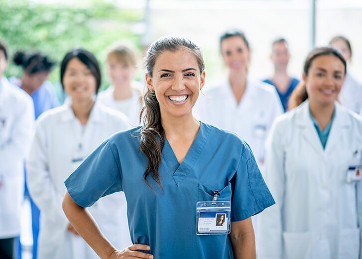 A female medical student of Hispanic ethnicity smiles for the camera as she stands in front of her group of medical professionals in the background.